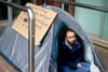 A man looks out from a black tent, pitched between two railings.