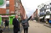 Two men stand on the corner of a city centre street. One is pointing up at something opposite.