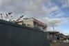 High hoarding is in the foreground and looming over is a boxy shopping centre, below a blue and cloudy sky.