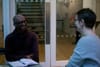 A man, with an open notepad in front of him, sits in conversation with a woman, who has her back to the camera.