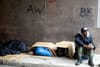 A man sits on the ground, with his knees pulled up to his chest and gazes to the right. Next to him in a laneway are cardboar