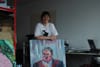 A woman in a white t-shirt stands behind a big portrait of a man, leaning her arms on the top edge.