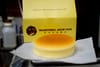 A round yellow cake sits with a golden top sits in front of a yellow takeaway box.